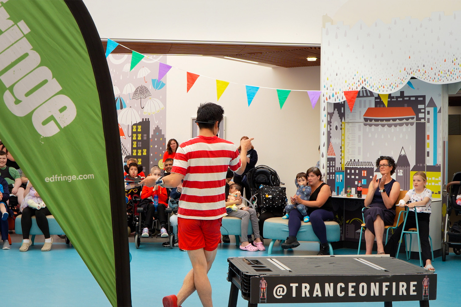 A Fringe artist in a stripey tshirt performs at the Edinburgh Children's Hospital; on a platform is the phrase '@TRANCEONFIRE'.